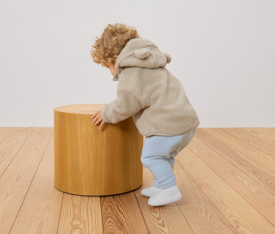Ein Kleinkind mit lockigem Haar und beigefarbener Teddyjacke mit Kapuze und Ohren stützt sich auf einen runden Holzhocker.