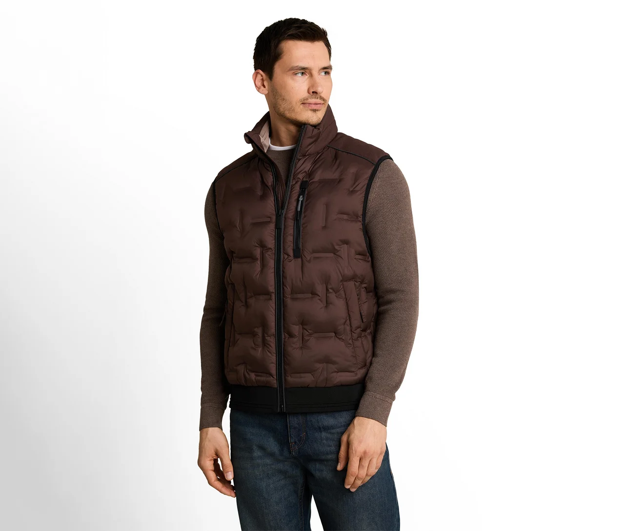 Ein Mann trägt eine braune Steppweste über einem braunen Langarmshirt und blauen Jeans und blickt nach rechts vor weißem Hintergrund.