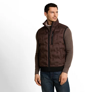 Ein Mann trägt eine braune Steppweste über einem braunen Langarmshirt und blauen Jeans und blickt nach rechts vor weißem Hintergrund.