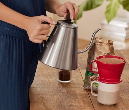Eine Person gießt Wasser aus einem silbernen Schwanenhalskessel in einen roten Kaffee Handfilter, Größe 101, der auf einer Tasse steht. Daneben liegen eine Kaffeebohnenpackung und gemahlener Kaffee.