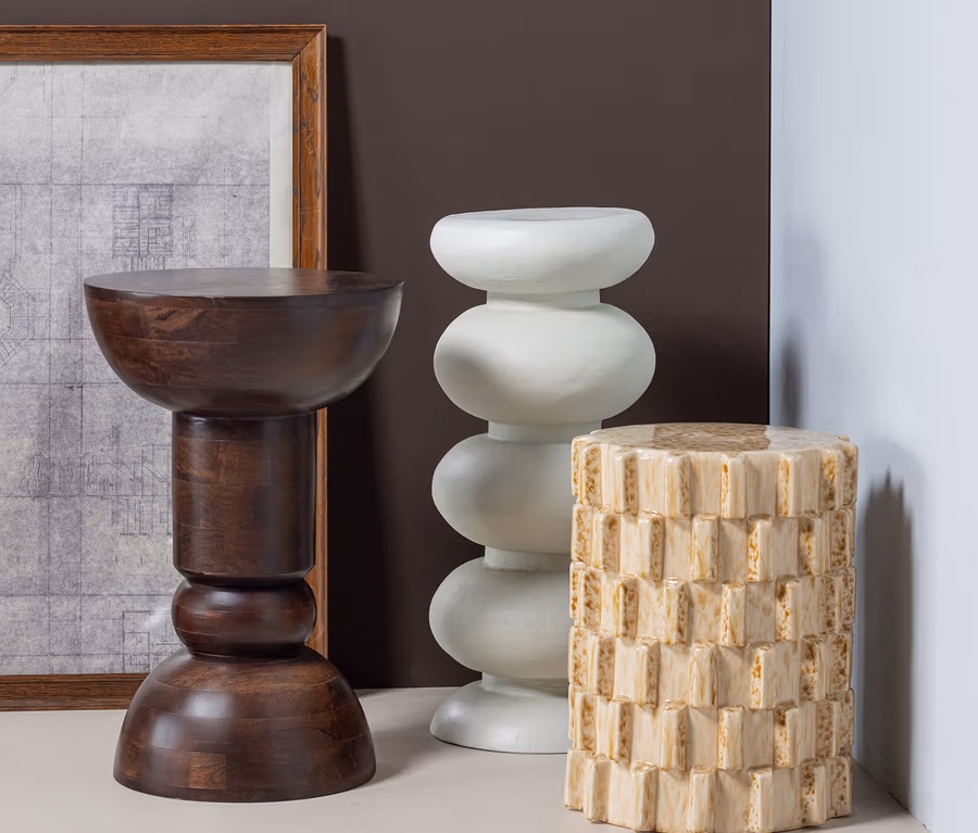 Drei Beistelltische in verschiedenen Designs: ein dunkelbrauner Holzhocker, ein cremefarbener Hocker mit drei bauchigen Segmenten und ein beigefarbener Zylinderhocker mit rechteckiger Textur.