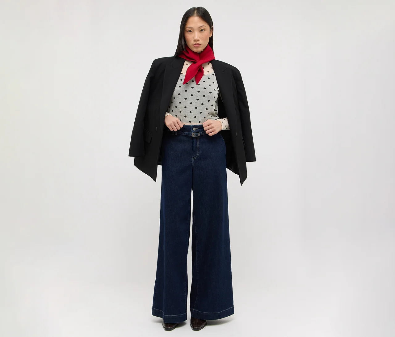 Eine Frau steht da und trägt einen schwarzen Blazer über den Schultern, ein helles gepunktetes Oberteil, dunkelblaue weite Jeans und einen roten Schal um den Hals.