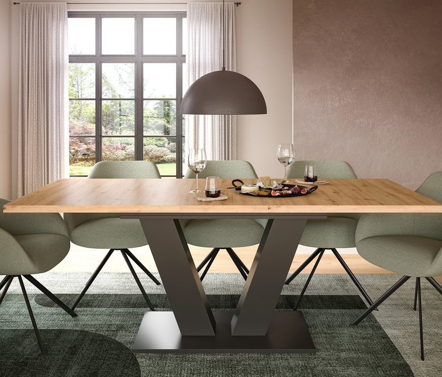 Ein Esszimmer mit einem Holztisch und sechs grünen Stühlen, über dem Tisch hängt eine dunkle Pendelleuchte. Auf dem Tisch stehen Käse, Feigen und Weingläser. Im Hintergrund ist ein großes Fenster mit Blick auf einen Garten zu sehen.