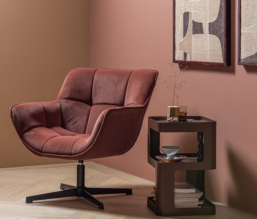 Ein roter, gesteppter Sessel mit schwarzem Drehfuß steht in einer Ecke mit rosa und braunen Wänden. Daneben ein brauner, mehrstufiger Beistelltisch mit einem Glas, einer Vase und Büchern. An der Wand hängen zwei gerahmte, abstrakte Bilder.