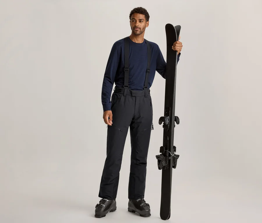 Ein Mann mit dunklem Lockenhaar und hellem Bart steht da, trägt ein dunkelblaues Langarmshirt, schwarze Skihosen mit Hosenträgern und schwarze Skischuhe. Er hält ein Paar schwarze Skier aufrecht in seiner rechten Hand.