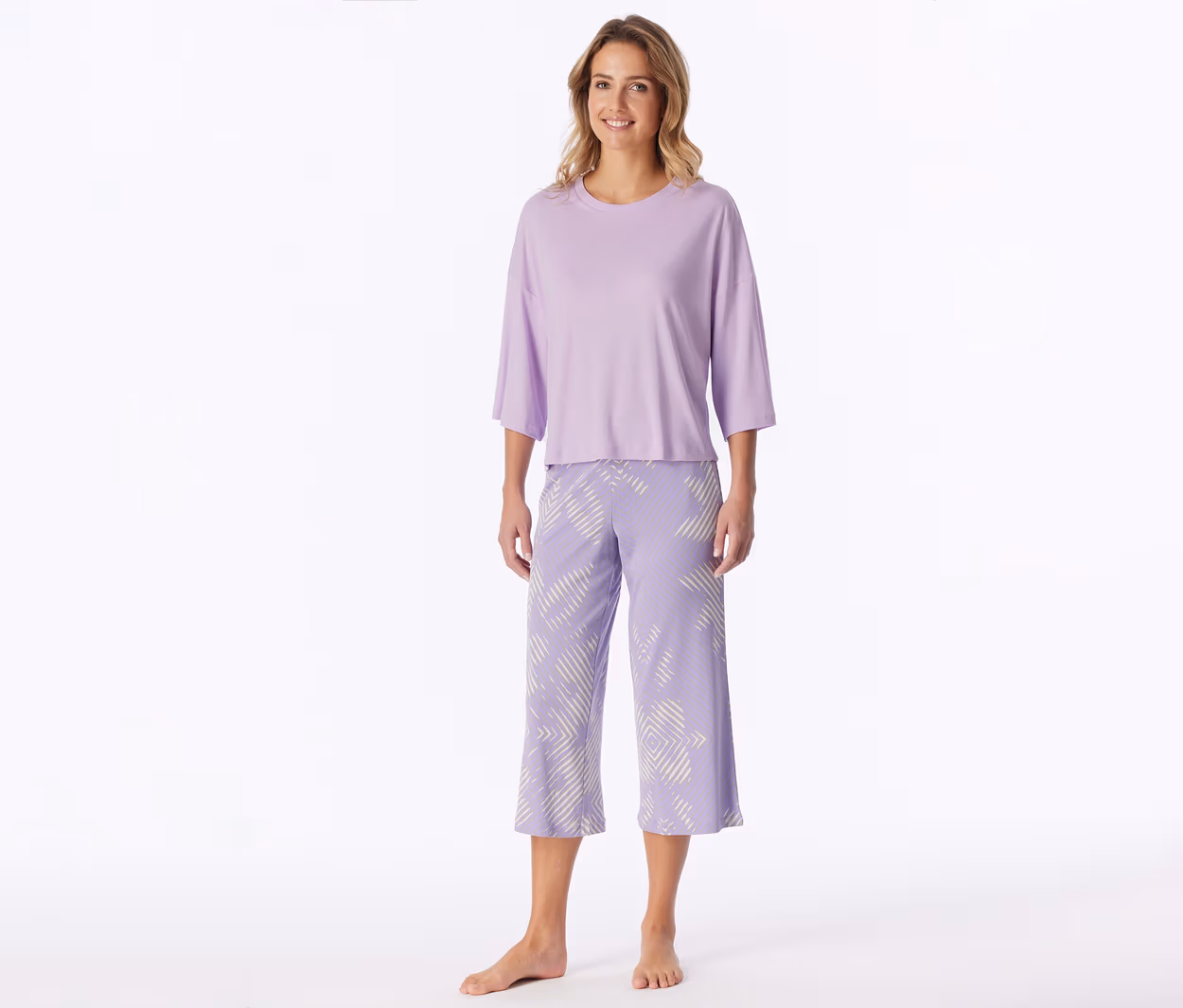 Lächelnde Frau steht barfuß und trägt ein hellviolettes T-Shirt mit Dreiviertelärmeln und passende Caprihosen mit weißem geometrischem Muster.