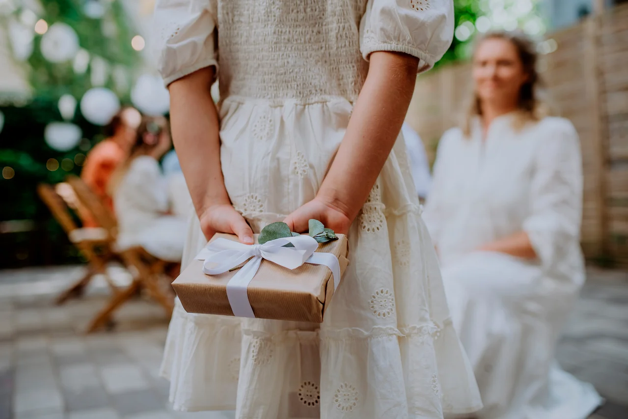 Kind in weißem Lochstickerei-Kleid hält ein braunes Geschenk mit weißer Schleife und Eukalyptusblättern hinter dem Rücken, mit unscharfen Personen im Hintergrund.
