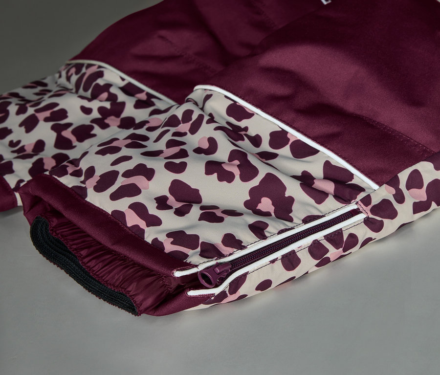 Nahaufnahme eines Kleidungsstücks aus bordeauxrotem und Leopardenmuster-Stoff. Das Leopardenmuster, beige mit dunkelrosa Flecken, wird durch einen weißen Streifen vom bordeauxroten Stoff getrennt. Ein bordeauxroter Reißverschluss und ein elastischer Bund sind am Leopardenmuster-Bereich sichtbar.