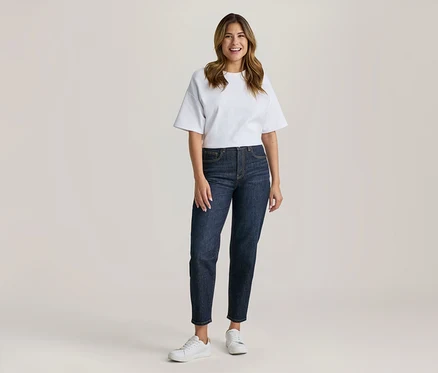 Frau steht lächelnd in weißem T-Shirt, dunkler Jeans und weißen Turnschuhen.