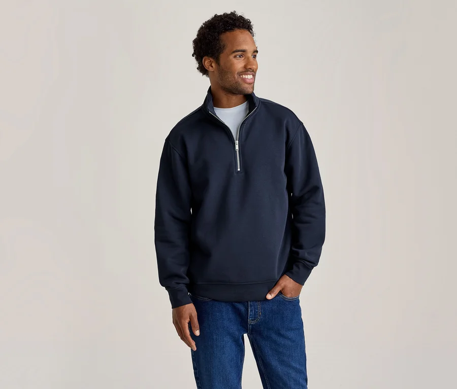Mann in einem marineblauen Quarter-Zip-Sweatshirt und blauen Jeans, lächelt und blickt nach rechts, eine Hand in der Hosentasche.
