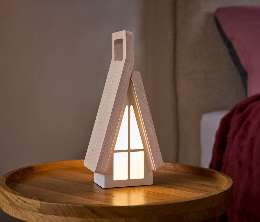 Eine hausförmige Lampe mit hellen Holzseiten und leuchtenden weißen Fenstern steht auf einem runden Holztisch. Im Hintergrund ist ein Bett mit einem rosa Kissen und einer roten Decke zu sehen.