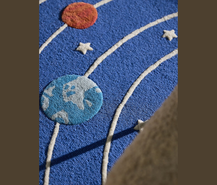 Blauer Teppich mit Weltraum-Motiv, darauf ein orangefarbener und ein blau-grauer Planet, weiße Sterne und weiße Umlaufbahnen. Rechts ein beiges Objekt.