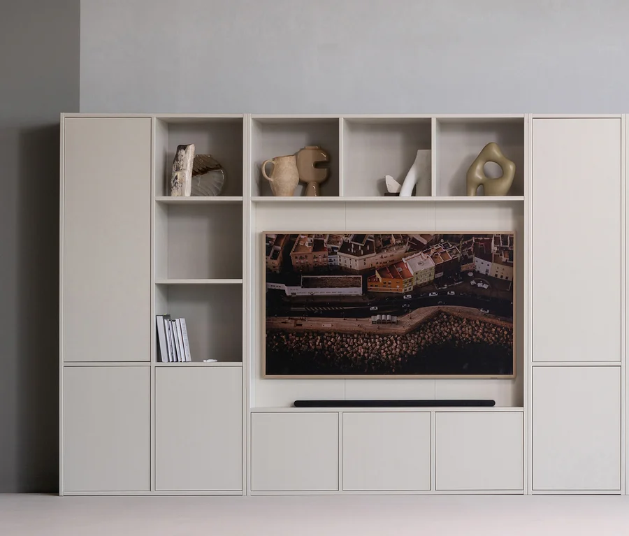 Helles, modulares Wandregal mit einem Fernseher, der eine Luftaufnahme einer Küstenstadt zeigt. Über dem Fernseher sind offene Fächer mit dekorativen Objekten, darunter Vasen und Skulpturen. Unter dem Fernseher befindet sich eine Soundbar.