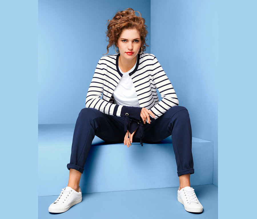 Eine Frau sitzt vor einem blauen Hintergrund und trägt eine Feinstrickjacke mit Volantärmeln, ein weißes T-Shirt, eine dunkelblaue Hose und weiße Turnschuhe.