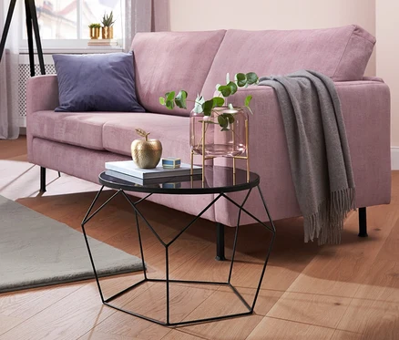 Ein Wohnzimmer mit einem rosa Max-Winzer®-Sofa 2,5-Sitzer »Cornelius«, einem Beistelltisch, einem Hochflorteppich, einem Hochglanz-Sideboard und einer Metall-Stehleuchte »Dreibein«.