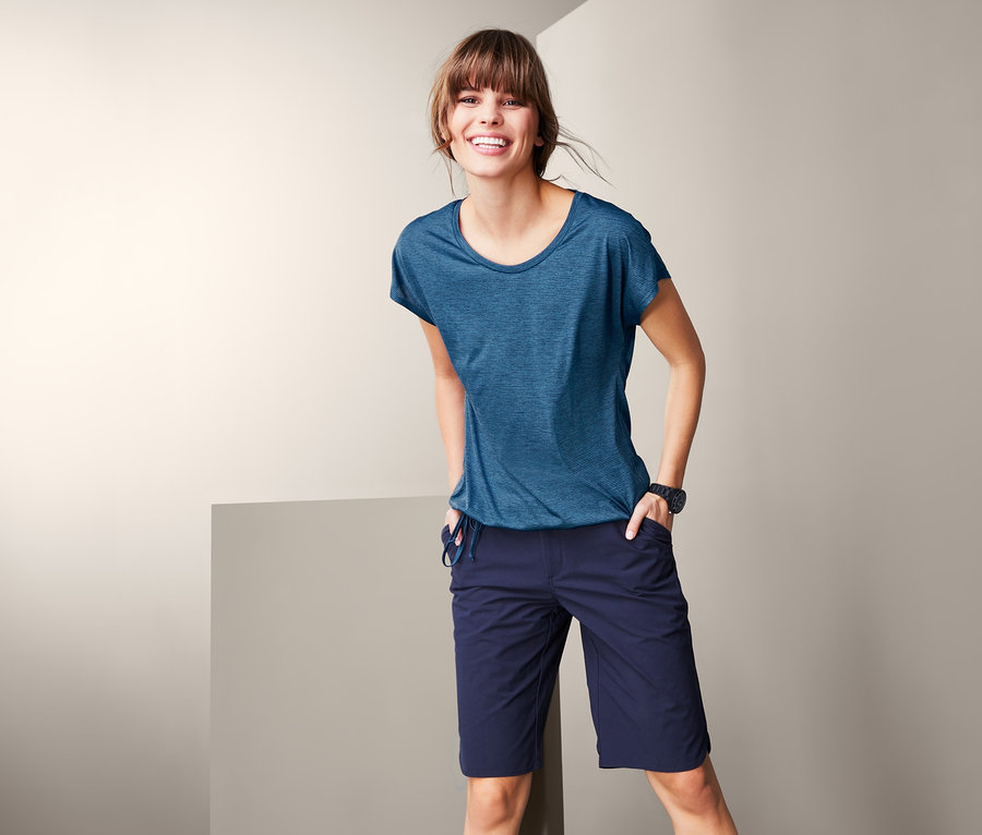 Eine lächelnde Frau posiert in einem blauen Funktionsshirt und einer dunkelblauen Shorts.