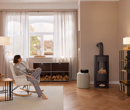 Eine Frau sitzt barfuß in einem cremefarbenen Schaukelstuhl mit Bouclé-Stoff. Links steht ein Beistelltisch mit Metallstreben, im Hintergrund ein Sideboard Metall »CN3« mit Brennholz und rechts ein Standregal mit Metallstreben.