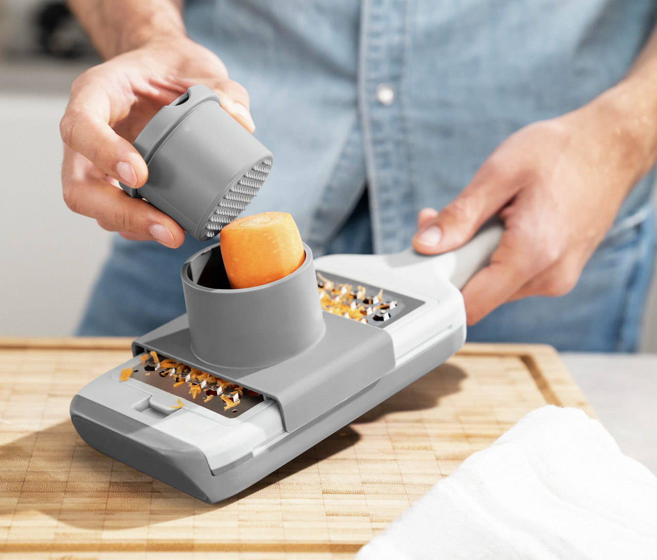 Eine Person reibt eine Karotte mit einer grau-weißen ZWILLING Z-Cut Multireibe. Eine Hand hält die Reibe, die andere schiebt die Karotte mit einem grauen Stößel hinein. Geriebene Karottenstücke liegen auf der Reibe, die auf einem Holzschneidebrett liegt.