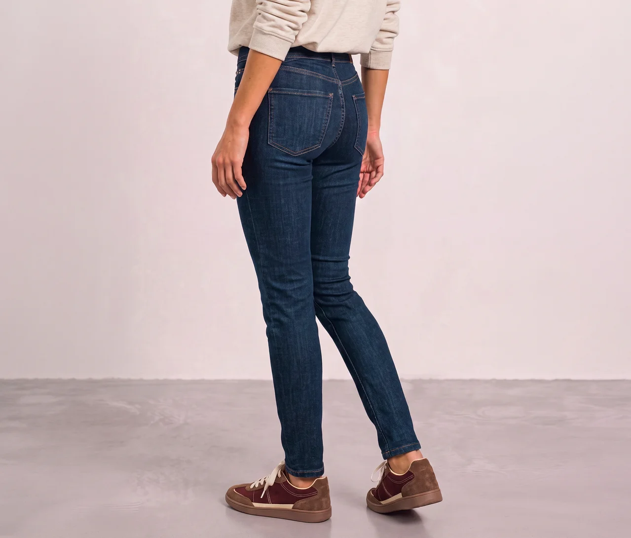 Eine Person von hinten trägt ein helles Oberteil, dunkelblaue Street One Jeans und braune Sneaker, während sie auf einem grauen Boden geht.