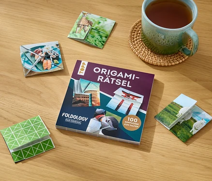 Auf einem Holztisch liegen das Buch »Origami-Rätsel«, eine Tasse Tee und gefaltete Origami-Figuren mit verschiedenen Motiven.
