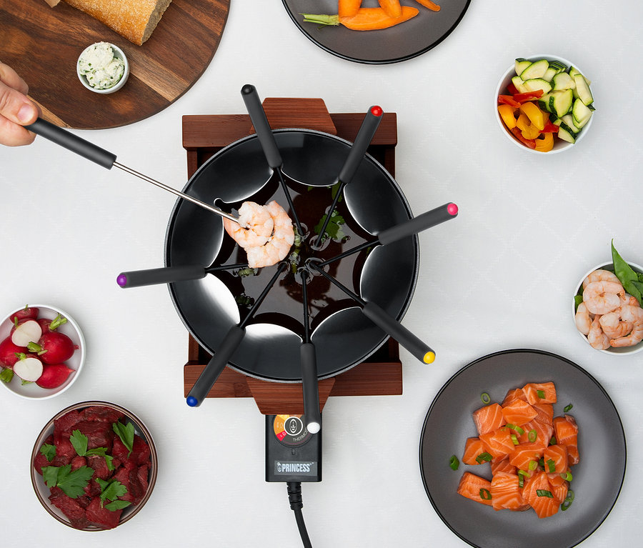 Draufsicht auf ein Fondue, bei dem eine Garnele auf einer Gabel aufgespießt ist, umgeben von Schüsseln mit Fleisch, Gemüse und Brot.