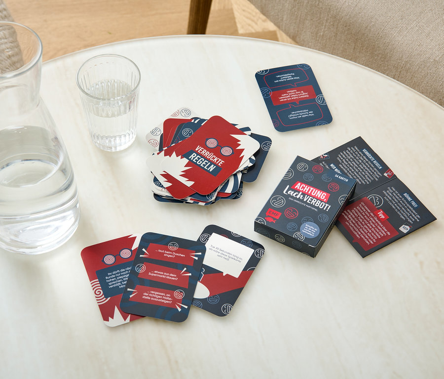 Auf einem Tisch liegen eine Glaskaraffe mit Wasser, ein Glas Wasser und Spielkarten des Kartenspiels »Achtung, Lachverbot!«.