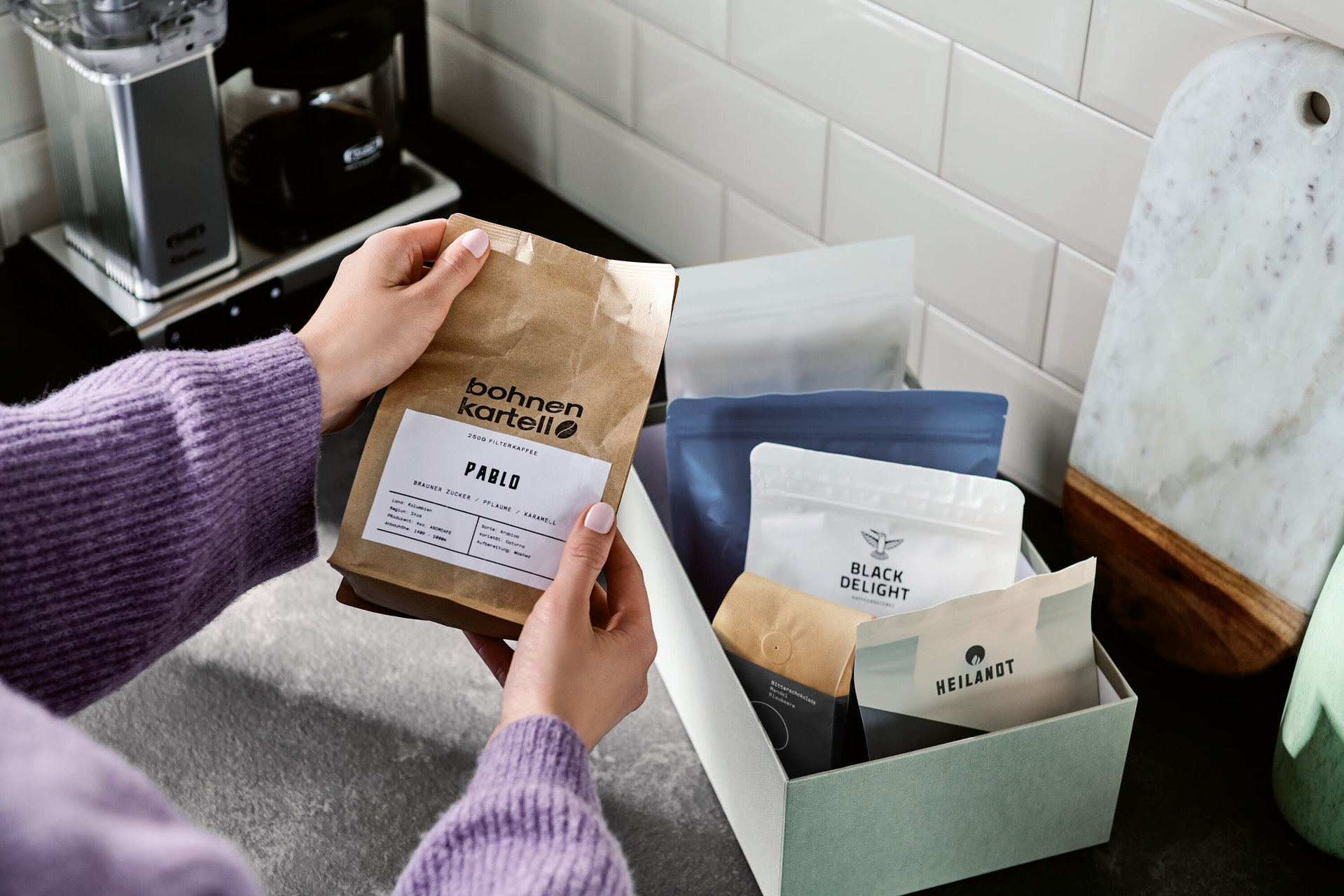 Frau hält eine Packung "bohnen kartell PABLO" Kaffee über einer Kiste mit anderen Kaffeepackungen.
