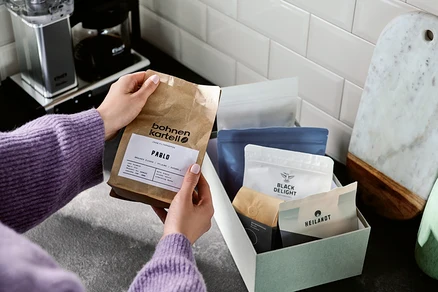 Frau hält eine Packung "bohnen kartell PABLO" Kaffee über einer Kiste mit anderen Kaffeepackungen.