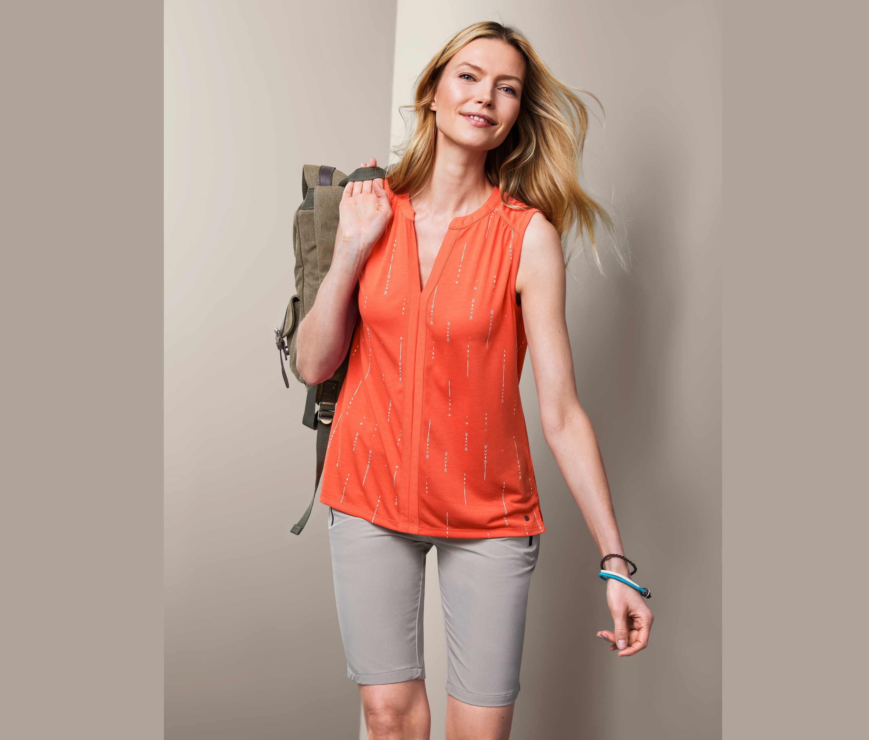 Frau mit blonden Haaren und Rucksack über der Schulter trägt ein orangefarbenes Funktionstop.