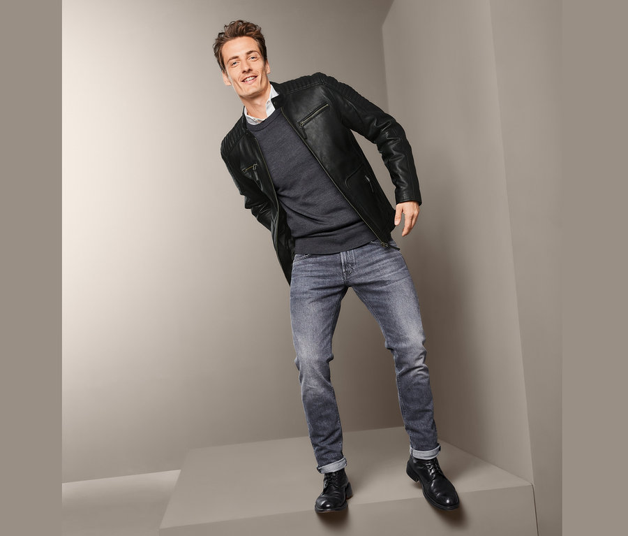 Ein Mann posiert in einer Lederjacke im Biker-Style, einem Pullover und Jeans.