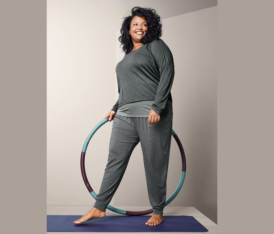 Frau steht auf einer Yogamatte, hält einen Hula-Hoop-Reifen und trägt ein 2-in-1-Sportshirt und eine Yogahose.