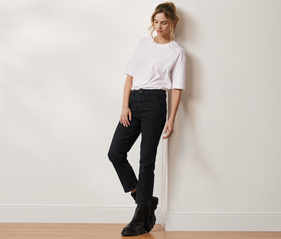 Eine Frau lehnt an einer Wand und trägt ein weißes T-Shirt, eine schwarze Cropped-Denim »Fit Alma« und schwarze Stiefel.