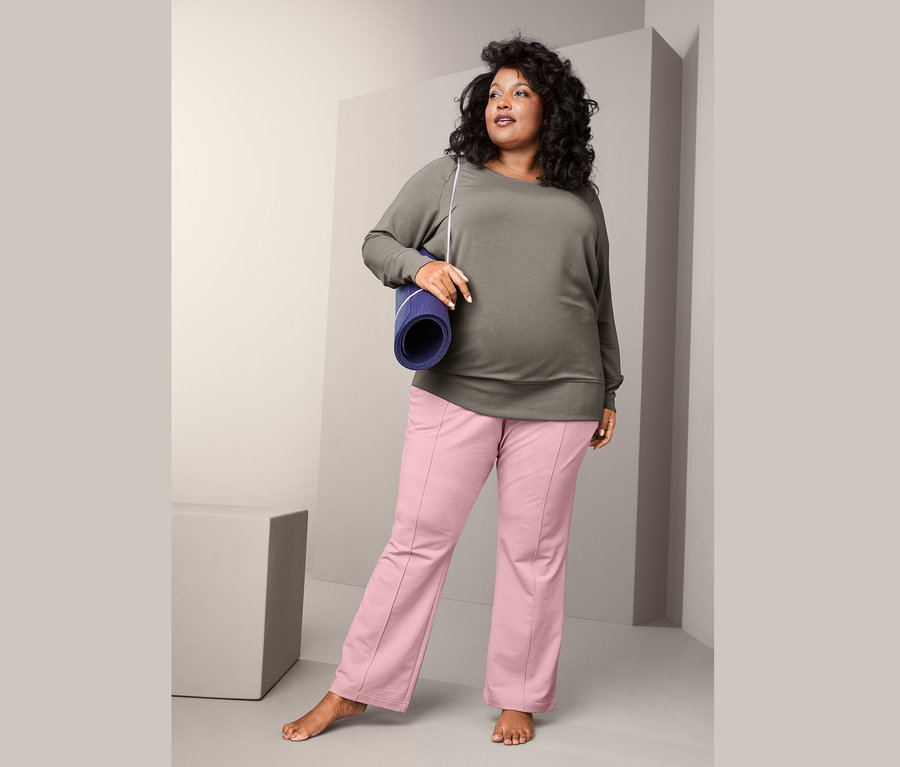 Frau trägt ein Yogasweatshirt und eine Sweathose und hält eine Yogamatte.