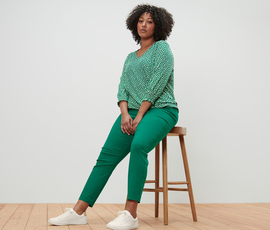 Eine Frau sitzt auf einem Hocker und trägt ein bedrucktes Blusenshirt in Grün und eine grüne Stretchhose.