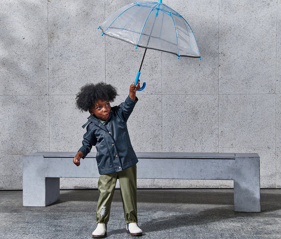 Junge hält einen transparenten Regenschirm mit blauer Umrandung über dem Kopf und trägt eine Regenjacke.