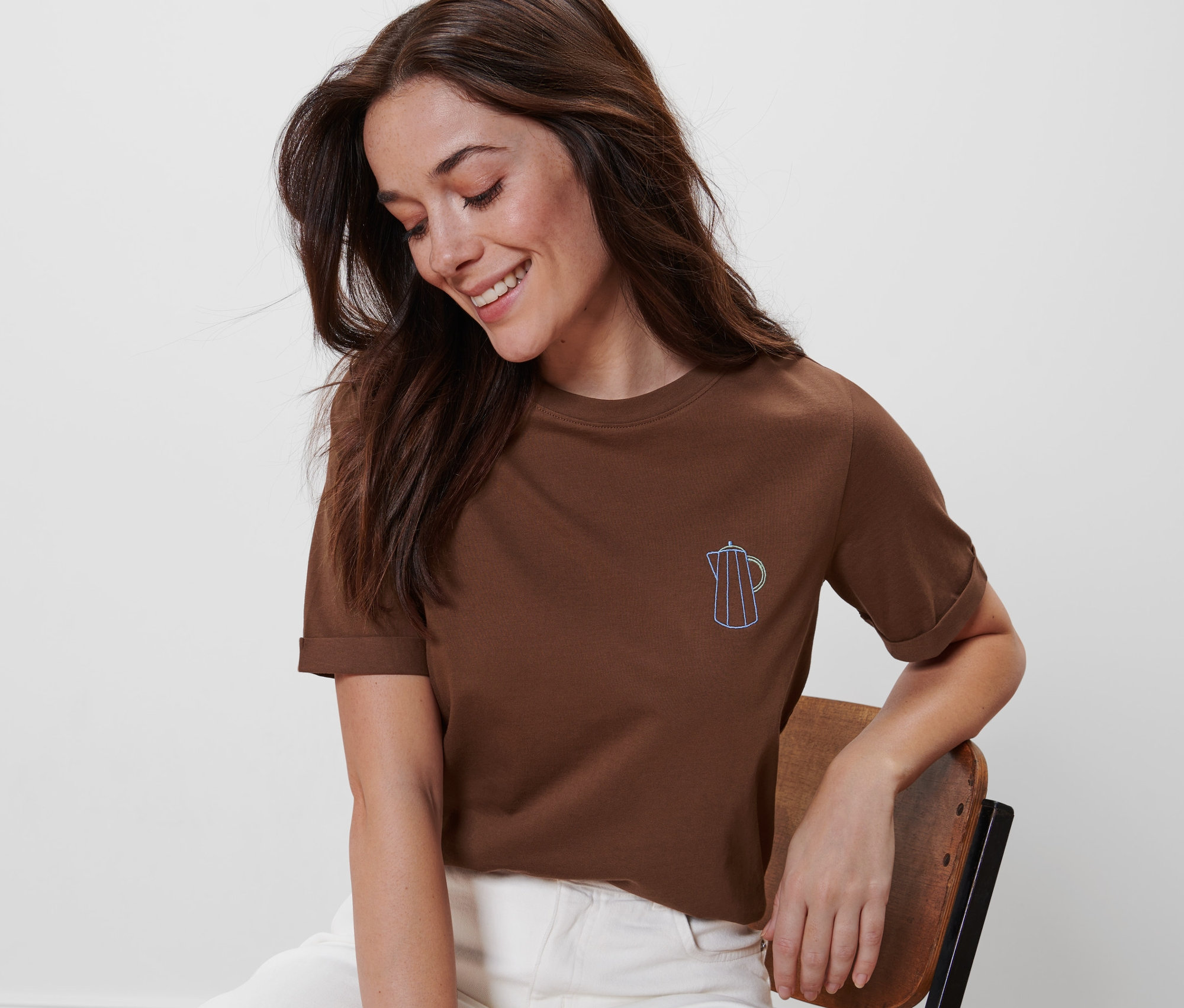 Frau sitzt auf einem Stuhl und trägt ein braunes T-Shirt mit Stickerei.