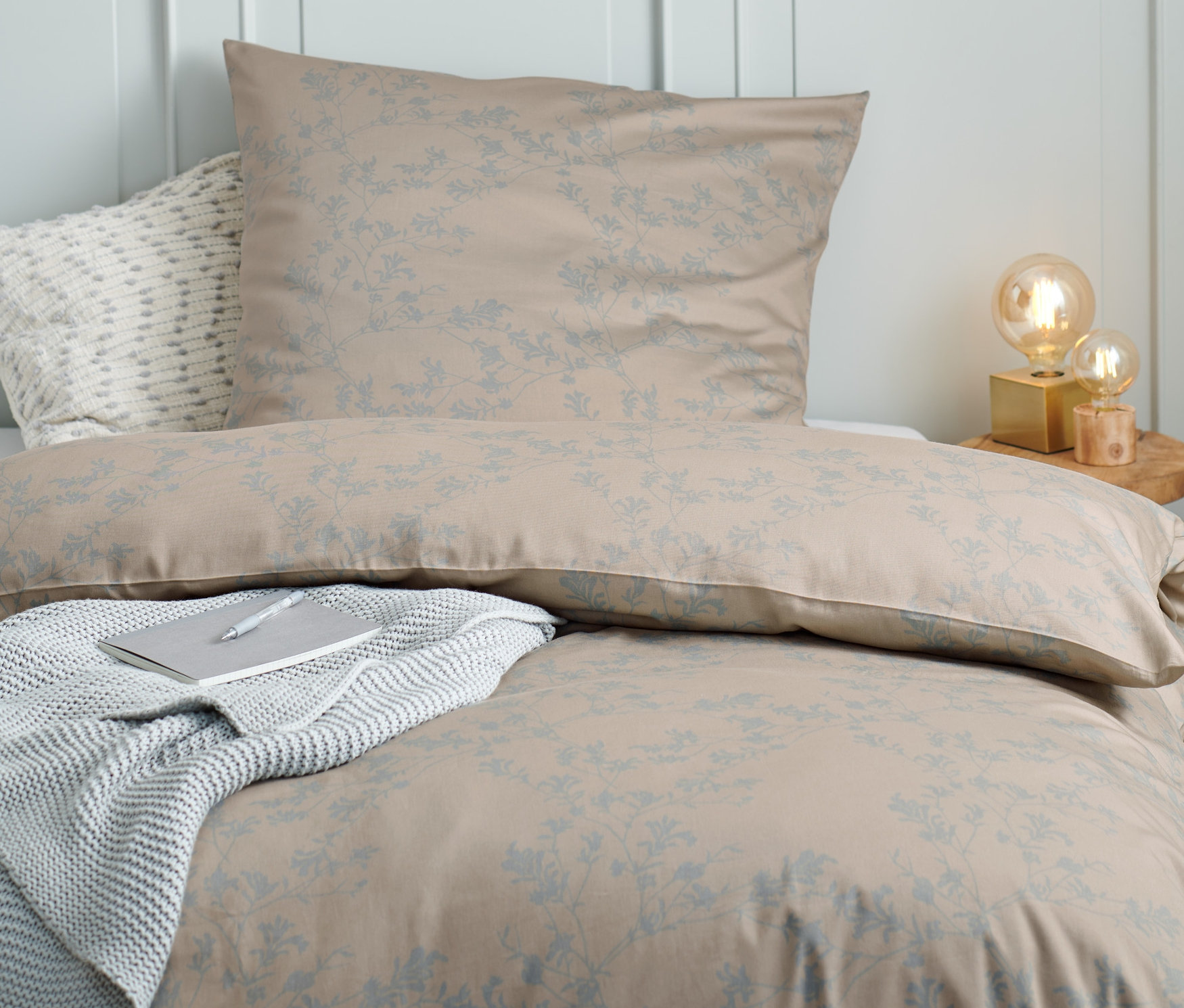 Beige-blaue Satin-Bettwäsche in Normalgröße und Übergröße auf einem Bett mit grauer Decke und Notizbuch.