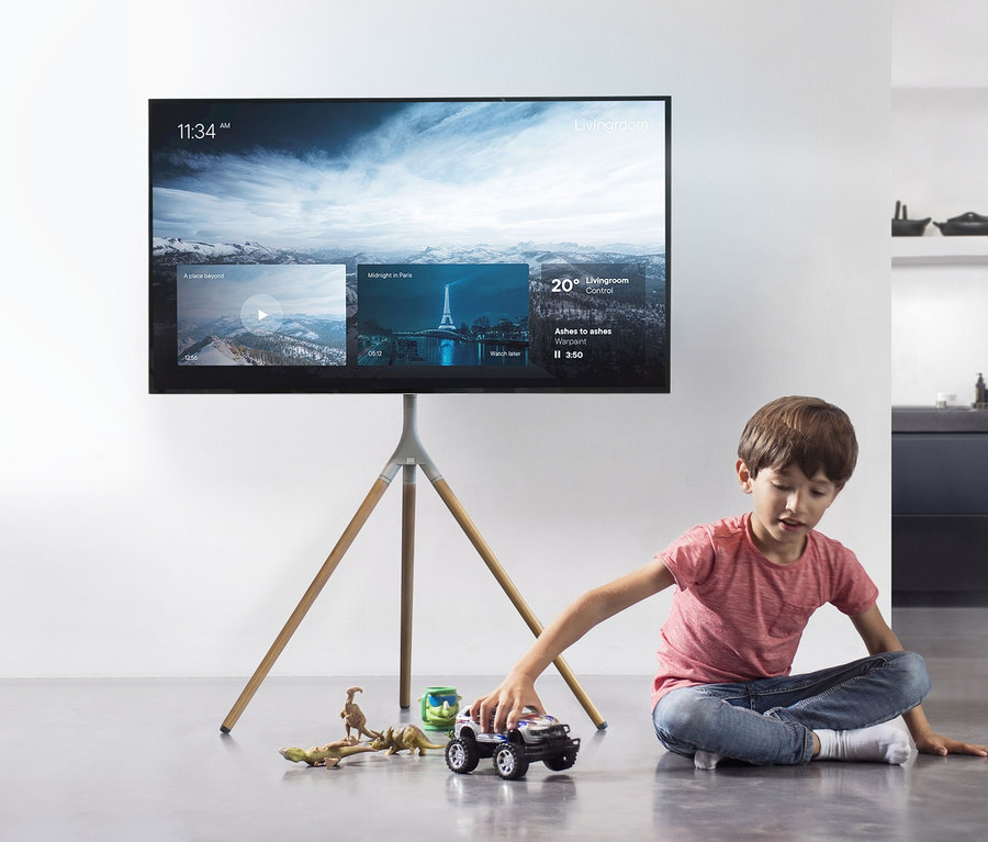 Ein Junge spielt mit einem Spielzeugauto auf dem Boden vor einem Fernseher, der auf einer ONE FOR ALL TRIPOD-TV-Halterung »WM7472« in Eiche und Silbergrau steht.