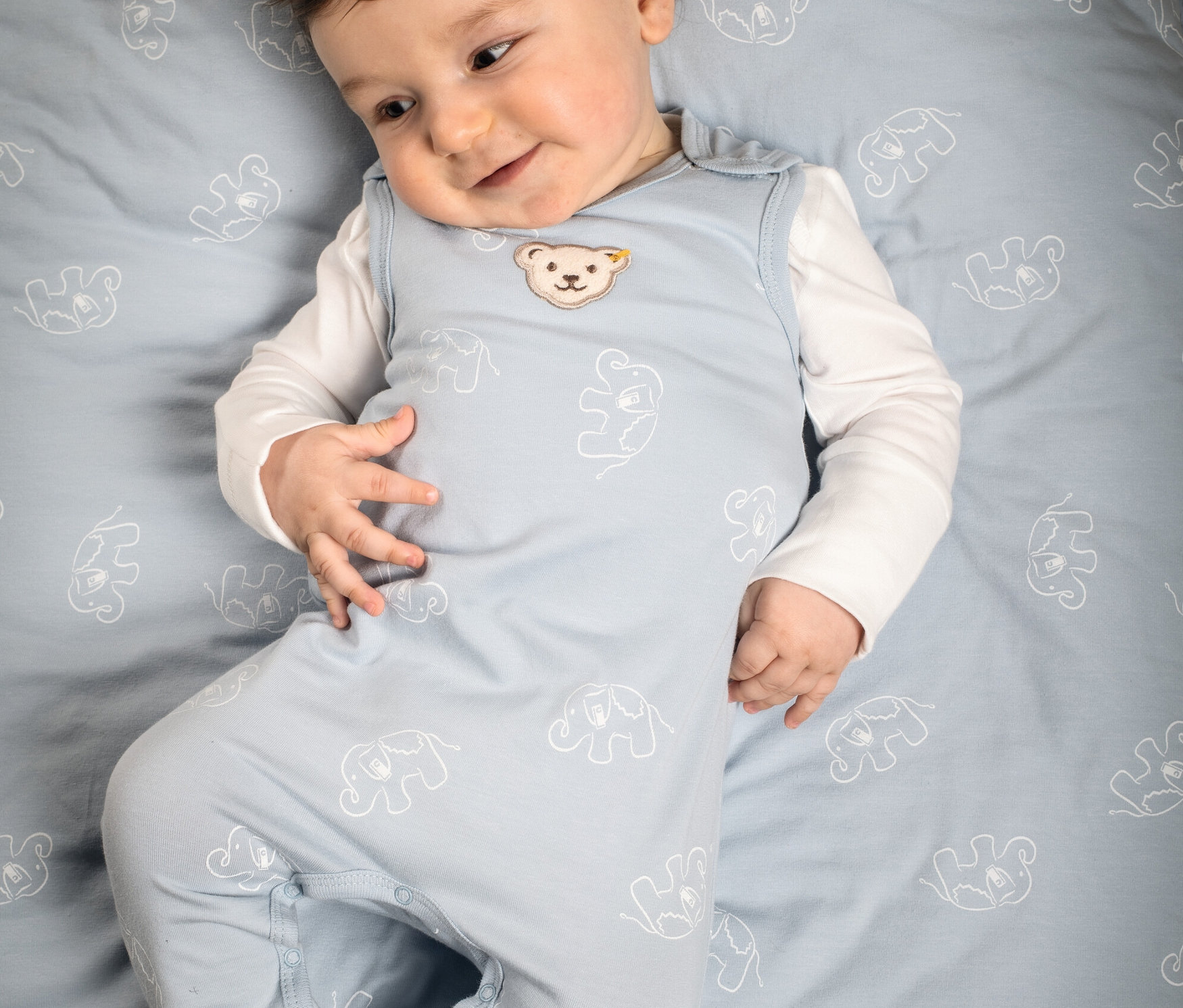 Baby liegt auf blauem Stoff mit Elefantenmuster und trägt einen hellblauen Steiff 2 Baby-Strampler.