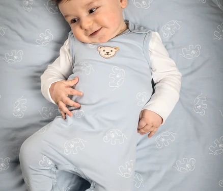 Ein lächelndes Baby liegt auf dem Rücken und trägt einen hellblauen Steiff Baby-Strampler mit Elefantenmuster und Bären-Logo über einem weißen Langarmshirt auf einem passenden Laken.