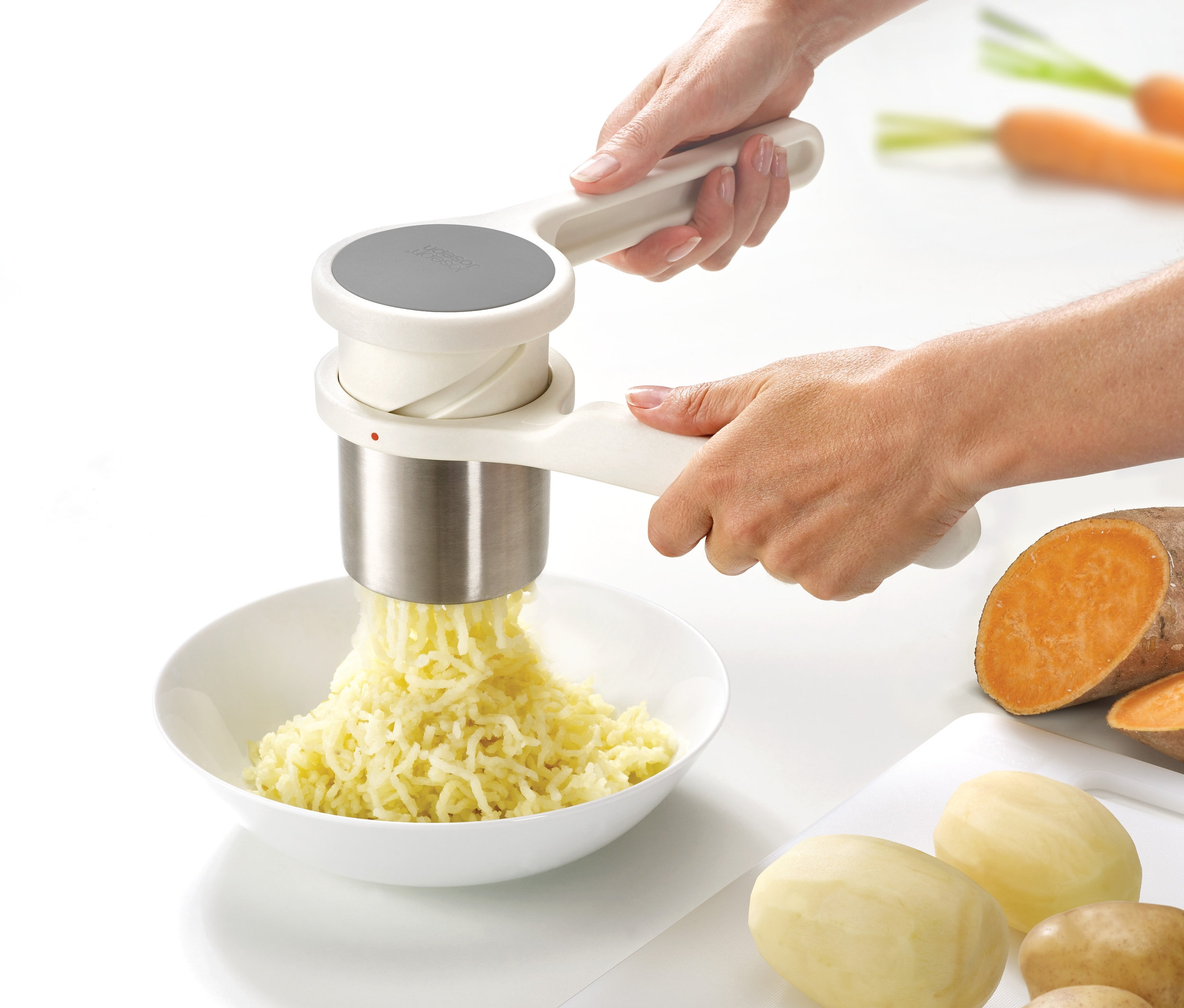 Person presst mit einer Joseph Joseph »Helix« Kartoffelpresse Kartoffeln in eine Schüssel. Auf dem Tisch liegen Kartoffeln, Süßkartoffeln und Karotten.