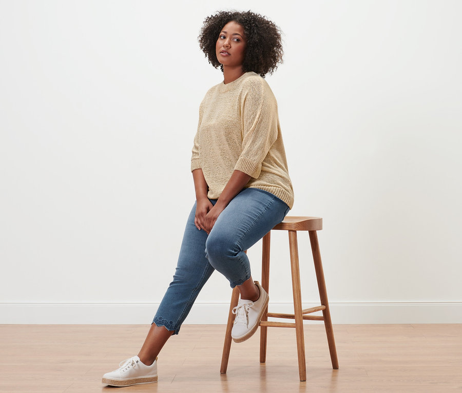 Frau sitzt auf einem Hocker und trägt eine 7/8-Treggings mit Lochstickerei und einen sommerlichen Strickpullover aus Bändchengarn.