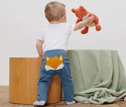 Kleinkind steht mit dem Rücken zur Kamera und hält einen Fuchs hoch. Es trägt 2 Babyhosen.