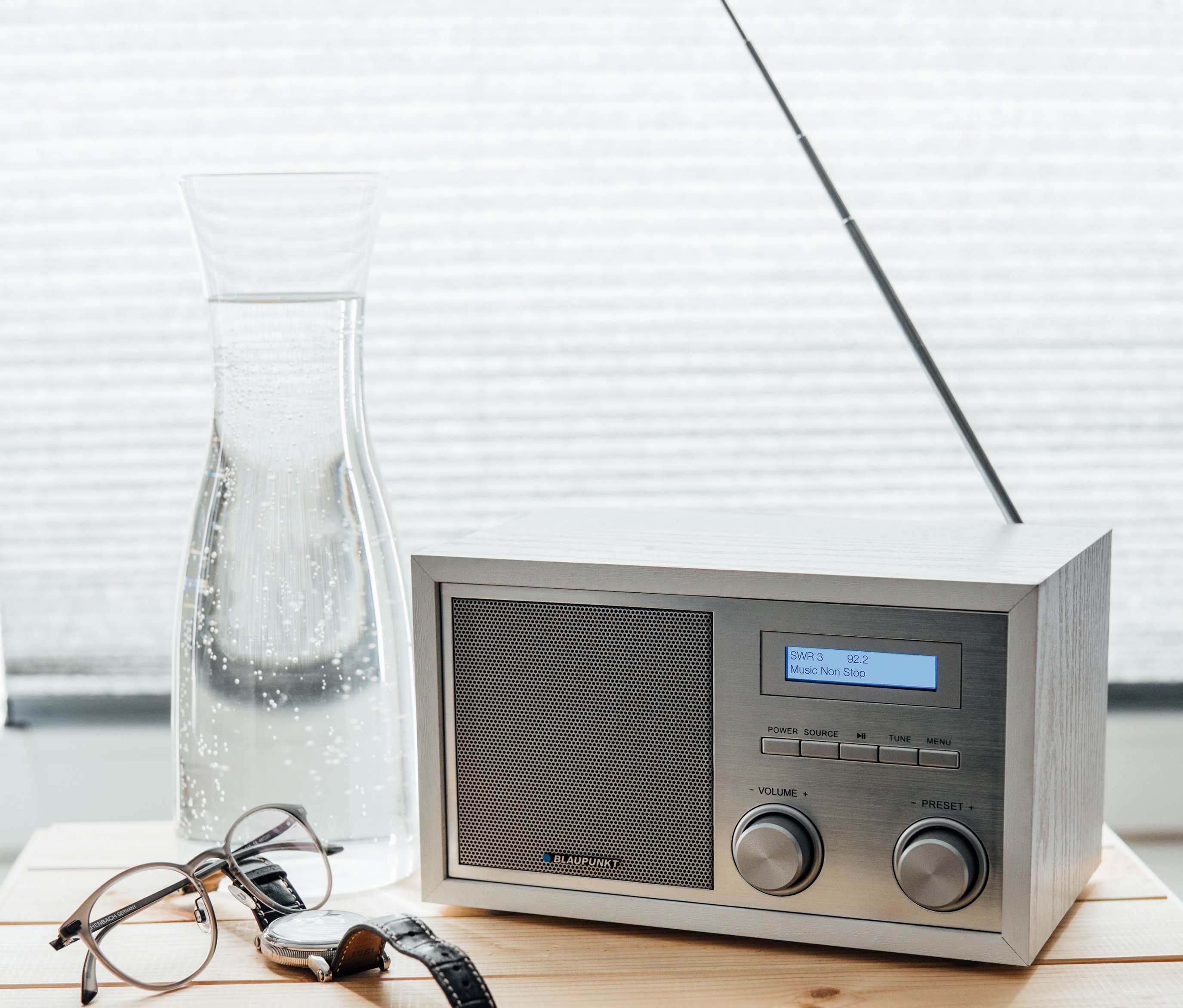 Eine Glaskaraffe mit Wasser, eine Brille und eine Uhr liegen neben einem weißen Blaupunkt DAB Radio »RXD180« auf einem Holztisch.