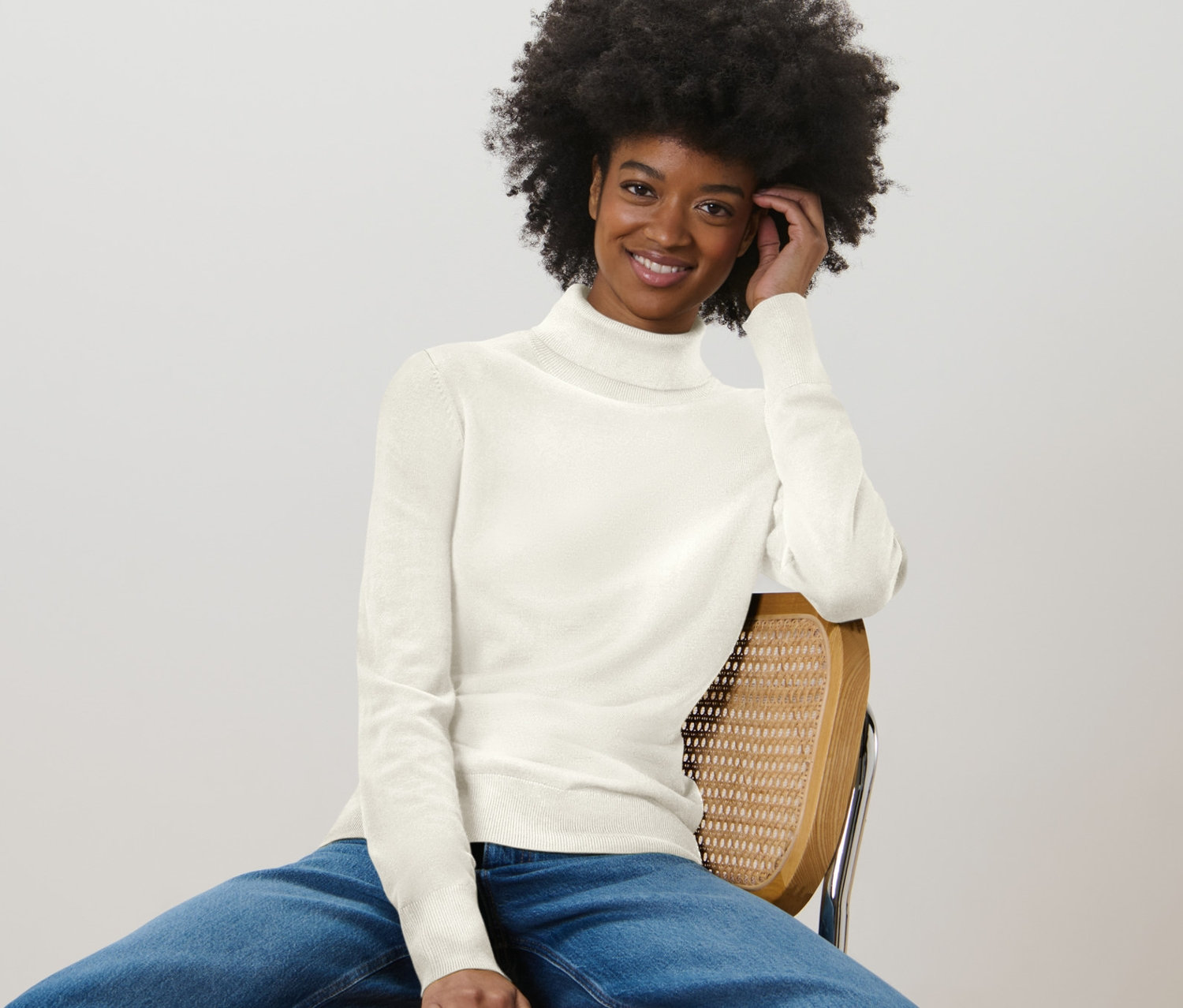 Frau sitzt auf einem Stuhl und trägt einen cremeweißen Feinstrick-Rollkragenpullover und Jeans.