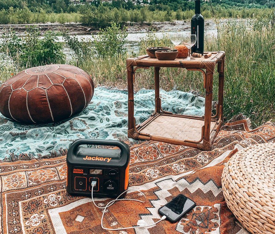 Bei einem Picknick am Fluss liegen ein brauner Puff und eine schwarze Ladestation Jackery Explorer 240 auf einer Decke, an die ein Mobiltelefon angeschlossen ist.