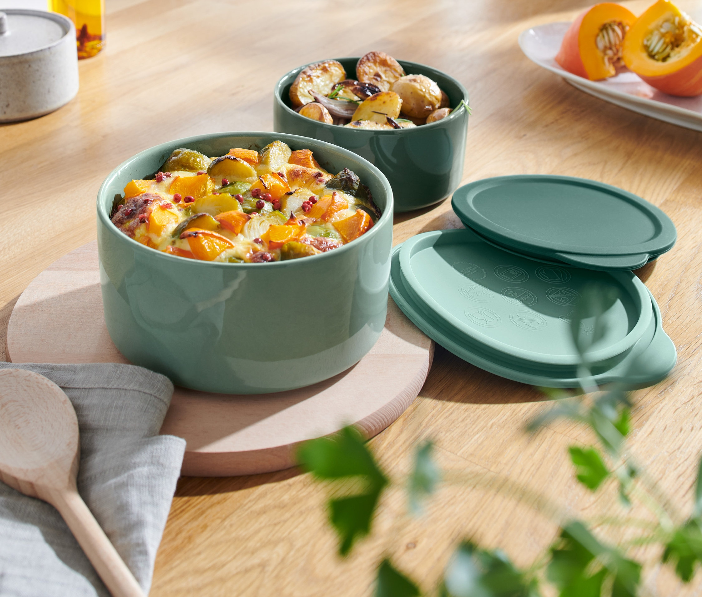 Zwei grüne Keramik-Auflaufformen mit Speisen stehen auf einem Holztisch.