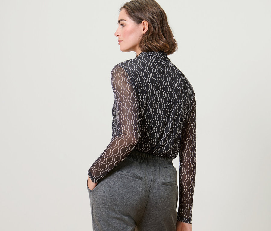 Frau steht mit dem Rücken zur Kamera und trägt ein bedrucktes Mesh-Rollkragenshirt und graue Hosen.