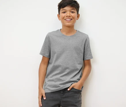 Ein Junge steht und trägt ein graues T-Shirt und Jeans. Er trägt 3 Kinder-T-Shirts in Schwarz, Grau und Weiß.