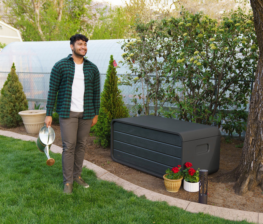 Mann steht im Garten neben einer grauen Aufbewahrungsbox (515 l) und hält eine Gießkanne.